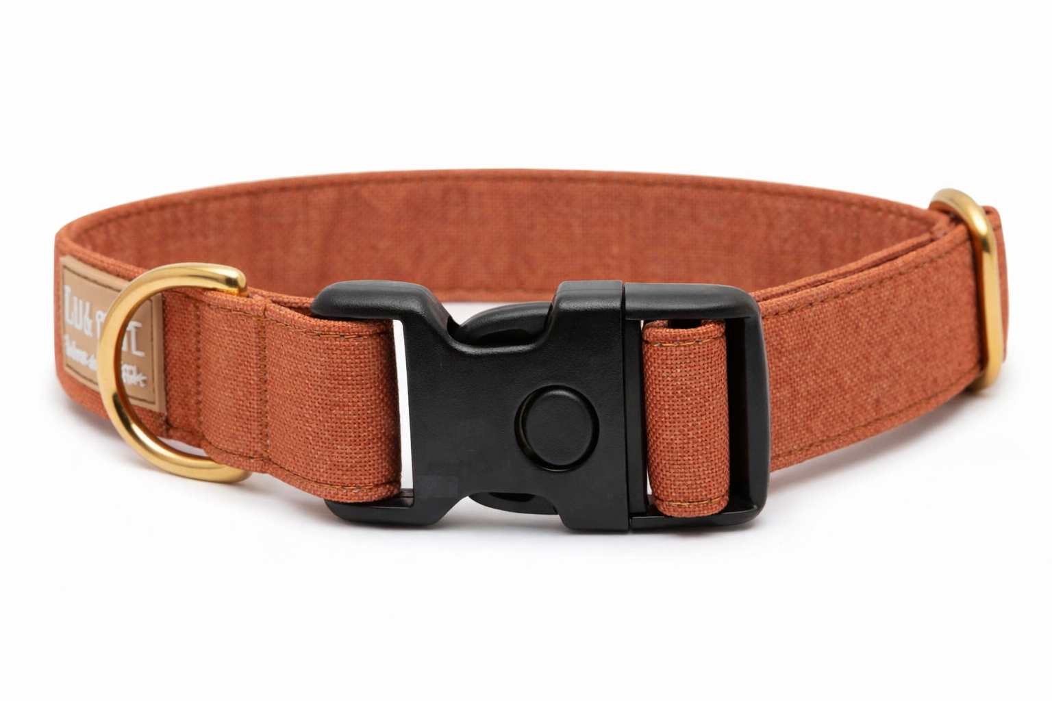 Hellbraunes Hundehalsband aus Stoff mit schwarzem Kunststoff-Schnellverschluss und goldenem Metallring zur Befestigung einer Leine. Das Halsband ist vor weißem Hintergrund fotografiert und zeigt eine breite, verstellbare Ausführung mit gepolsterter Innenseite. Auf einem kleinen Etikett am Ring ist ein Markenlogo zu erkennen.