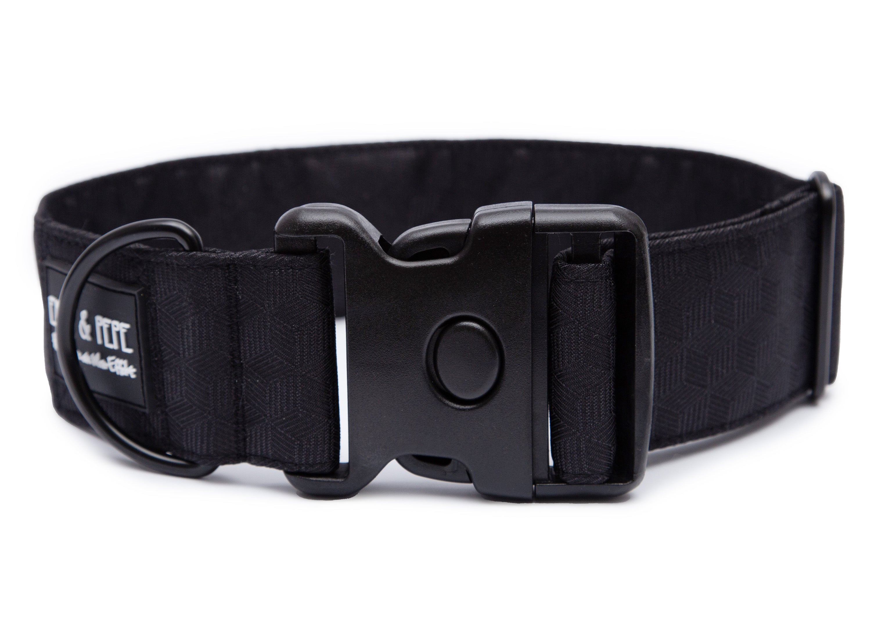 Schwarzes Hundehalsband aus Stoff mit schwarzem Kunststoff-Schnellverschluss und schwarzem Metallring zur Befestigung einer Leine. Das Halsband ist vor weißem Hintergrund fotografiert und zeigt eine breite, verstellbare Ausführung mit gepolsterter Innenseite. Auf einem kleinen Etikett am Ring ist ein Markenlogo zu erkennen.