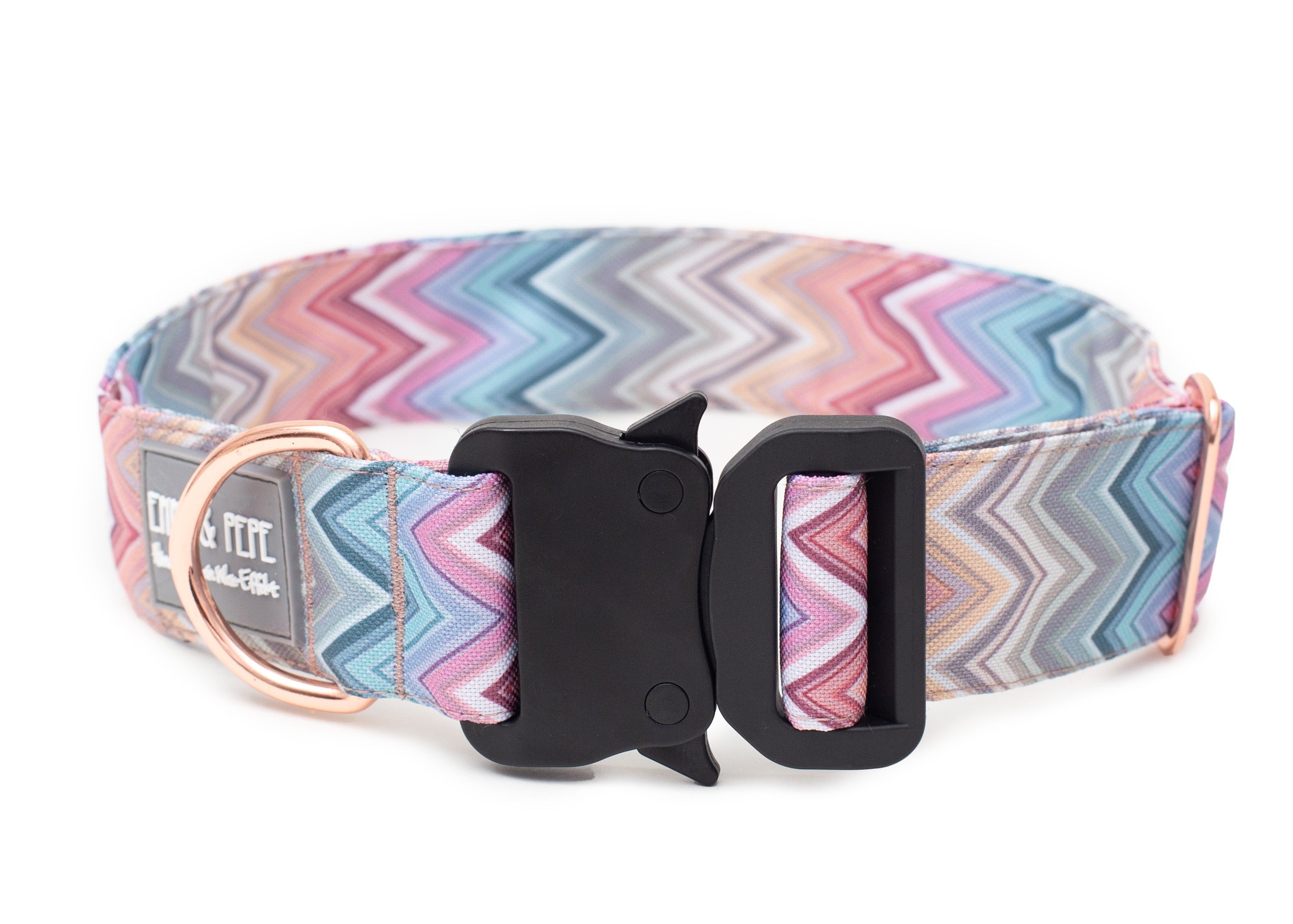 pastellfarbenes Zickzackmuster Hundehalsband aus Stoff mit schwarzem Kunststoff-Sicherheitsverschluss und rosegoldenem Metallring zur Befestigung einer Leine. Das Halsband ist vor weißem Hintergrund fotografiert und zeigt eine breite, verstellbare Ausführung mit gepolsterter Innenseite. Auf einem kleinen Etikett am Ring ist ein Markenlogo zu erkennen.