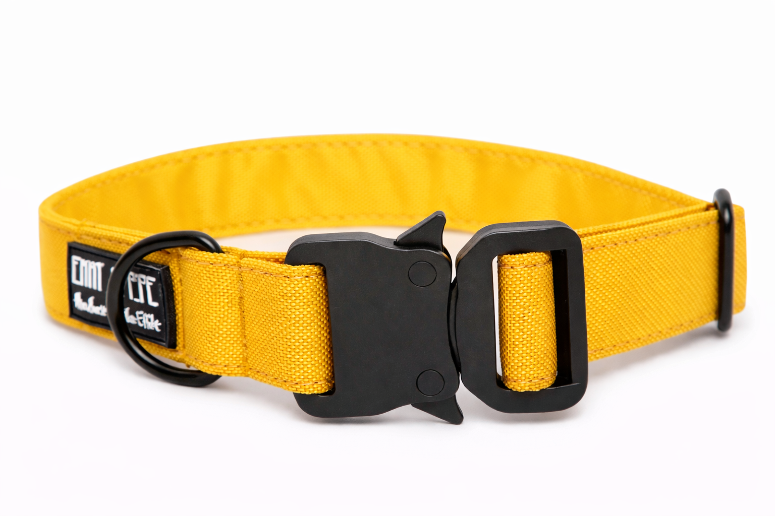 Gelbes Hundehalsband aus Stoff mit schwarzem Kunststoff-Sicherheitsverschluss und schwarzem Metallring zur Befestigung einer Leine. Das Halsband ist vor weißem Hintergrund fotografiert und zeigt eine breite, verstellbare Ausführung mit gepolsterter Innenseite. Auf einem kleinen Etikett am Ring ist ein Markenlogo zu erkennen.