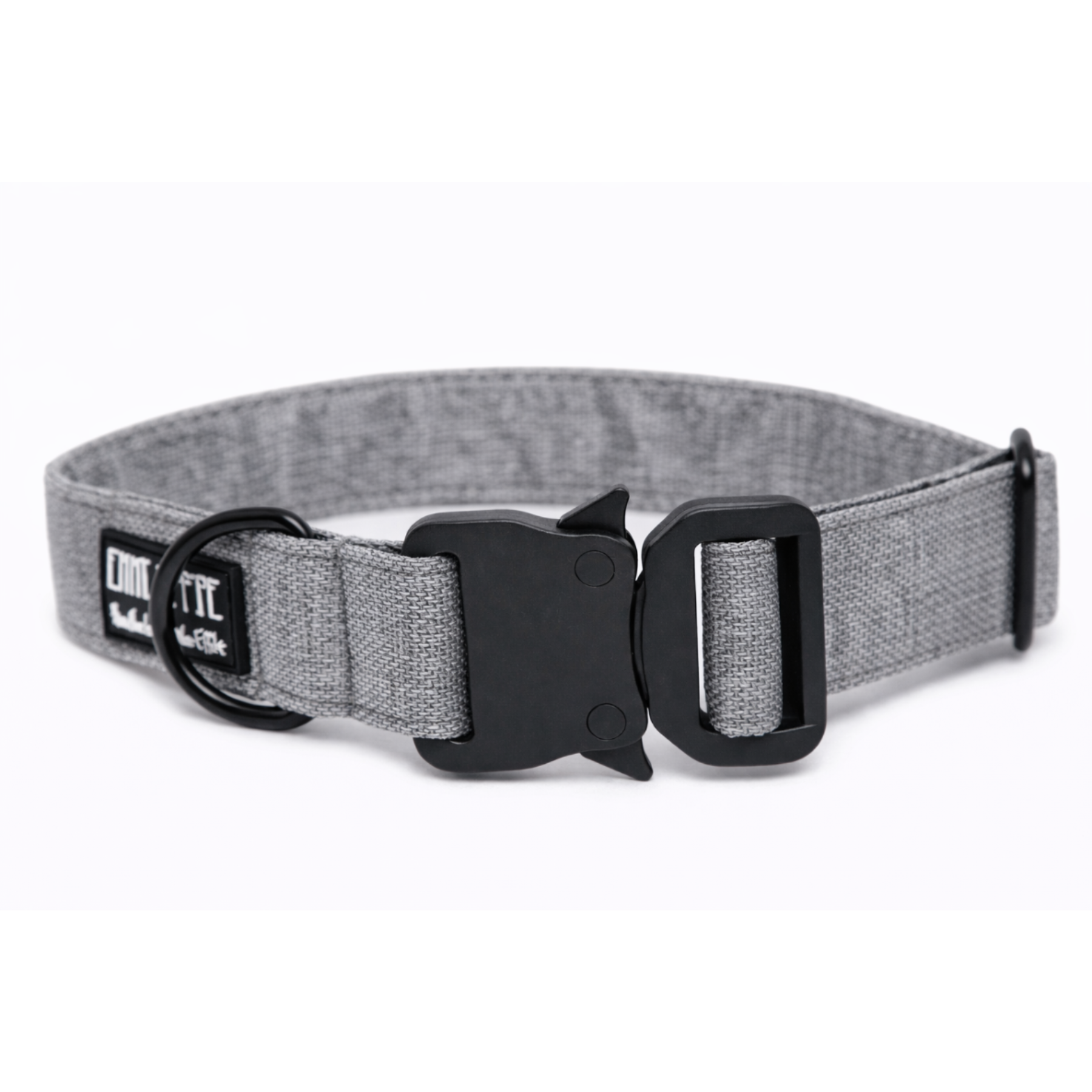Graues Hundehalsband aus Stoff mit schwarzem Kunststoff-Sicherheitsverschluss und schwarzem Metallring zur Befestigung einer Leine. Das Halsband ist vor weißem Hintergrund fotografiert und zeigt eine breite, verstellbare Ausführung mit gepolsterter Innenseite. Auf einem kleinen Etikett am Ring ist ein Markenlogo zu erkennen.