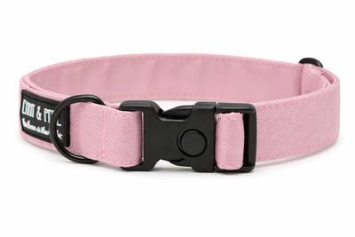 Rosafarbenes Hundehalsband aus Stoff mit schwarzem Kunststoff-Schnellverschluss und schwarzem Metallring zur Befestigung einer Leine. Das Halsband ist vor weißem Hintergrund fotografiert und zeigt eine breite, verstellbare Ausführung mit gepolsterter Innenseite. Auf einem kleinen Etikett am Ring ist ein Markenlogo zu erkennen.