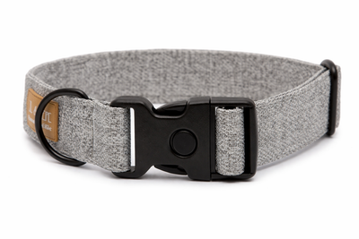 Graues Hundehalsband aus Stoff mit schwarzem Kunststoff-Schnellverschluss und schwarzem Metallring zur Befestigung einer Leine. Das Halsband ist vor weißem Hintergrund fotografiert und zeigt eine breite, verstellbare Ausführung mit gepolsterter Innenseite. Auf einem kleinen Etikett am Ring ist ein Markenlogo zu erkennen.