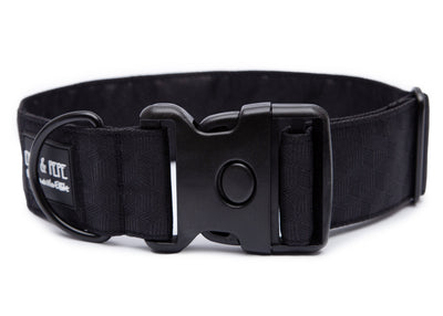 Schwarzes Hundehalsband aus Stoff mit schwarzem Kunststoff-Schnellverschluss und schwarzem Metallring zur Befestigung einer Leine. Das Halsband ist vor weißem Hintergrund fotografiert und zeigt eine breite, verstellbare Ausführung mit gepolsterter Innenseite. Auf einem kleinen Etikett am Ring ist ein Markenlogo zu erkennen.