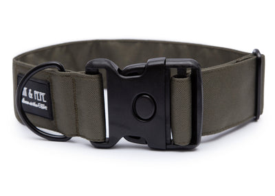 Olivefarbenes Hundehalsband aus Stoff mit schwarzem Kunststoff-Schnellverschluss und schwarzem Metallring zur Befestigung einer Leine. Das Halsband ist vor weißem Hintergrund fotografiert und zeigt eine breite, verstellbare Ausführung mit gepolsterter Innenseite. Auf einem kleinen Etikett am Ring ist ein Markenlogo zu erkennen.