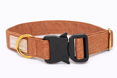 Hellbraunes Hundehalsband aus Stoff mit schwarzem Kunststoff-Sicherheitsverschluss und goldenem Metallring zur Befestigung einer Leine. Das Halsband ist vor weißem Hintergrund fotografiert und zeigt eine breite, verstellbare Ausführung mit gepolsterter Innenseite. Auf einem kleinen Etikett am Ring ist ein Markenlogo zu erkennen.
