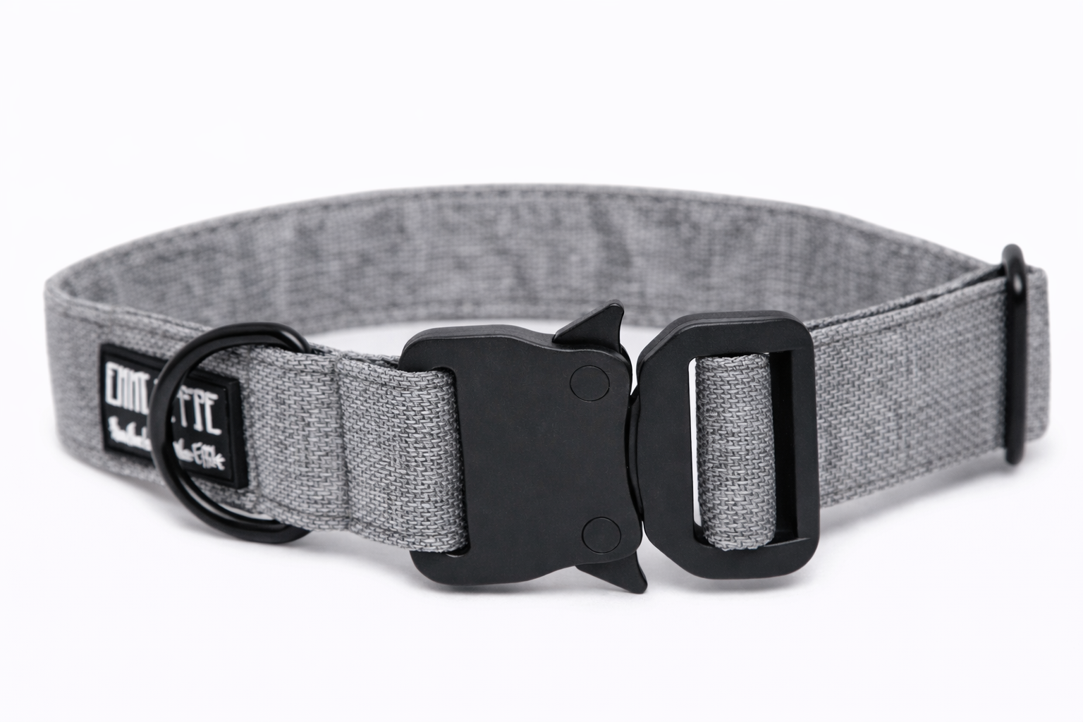 Graues Hundehalsband aus Stoff mit schwarzem Kunststoff-Sicherheitsverschluss und schwarzem Metallring zur Befestigung einer Leine. Das Halsband ist vor weißem Hintergrund fotografiert und zeigt eine breite, verstellbare Ausführung mit gepolsterter Innenseite. Auf einem kleinen Etikett am Ring ist ein Markenlogo zu erkennen.