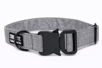 Graues Hundehalsband aus Stoff mit schwarzem Kunststoff-Sicherheitsverschluss und schwarzem Metallring zur Befestigung einer Leine. Das Halsband ist vor weißem Hintergrund fotografiert und zeigt eine breite, verstellbare Ausführung mit gepolsterter Innenseite. Auf einem kleinen Etikett am Ring ist ein Markenlogo zu erkennen.