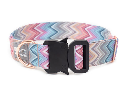 pastellfarbenes Zickzackmuster Hundehalsband aus Stoff mit schwarzem Kunststoff-Sicherheitsverschluss und rosegoldenem Metallring zur Befestigung einer Leine. Das Halsband ist vor weißem Hintergrund fotografiert und zeigt eine breite, verstellbare Ausführung mit gepolsterter Innenseite. Auf einem kleinen Etikett am Ring ist ein Markenlogo zu erkennen.