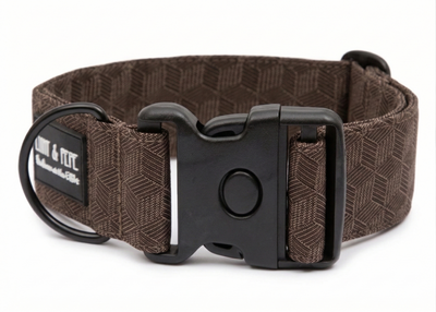 Braunes Hundehalsband aus Stoff mit schwarzem Kunststoff-Schnellverschluss und schwarzem Metallring zur Befestigung einer Leine. Das Halsband ist vor weißem Hintergrund fotografiert und zeigt eine breite, verstellbare Ausführung mit gepolsterter Innenseite. Auf einem kleinen Etikett am Ring ist ein Markenlogo zu erkennen.