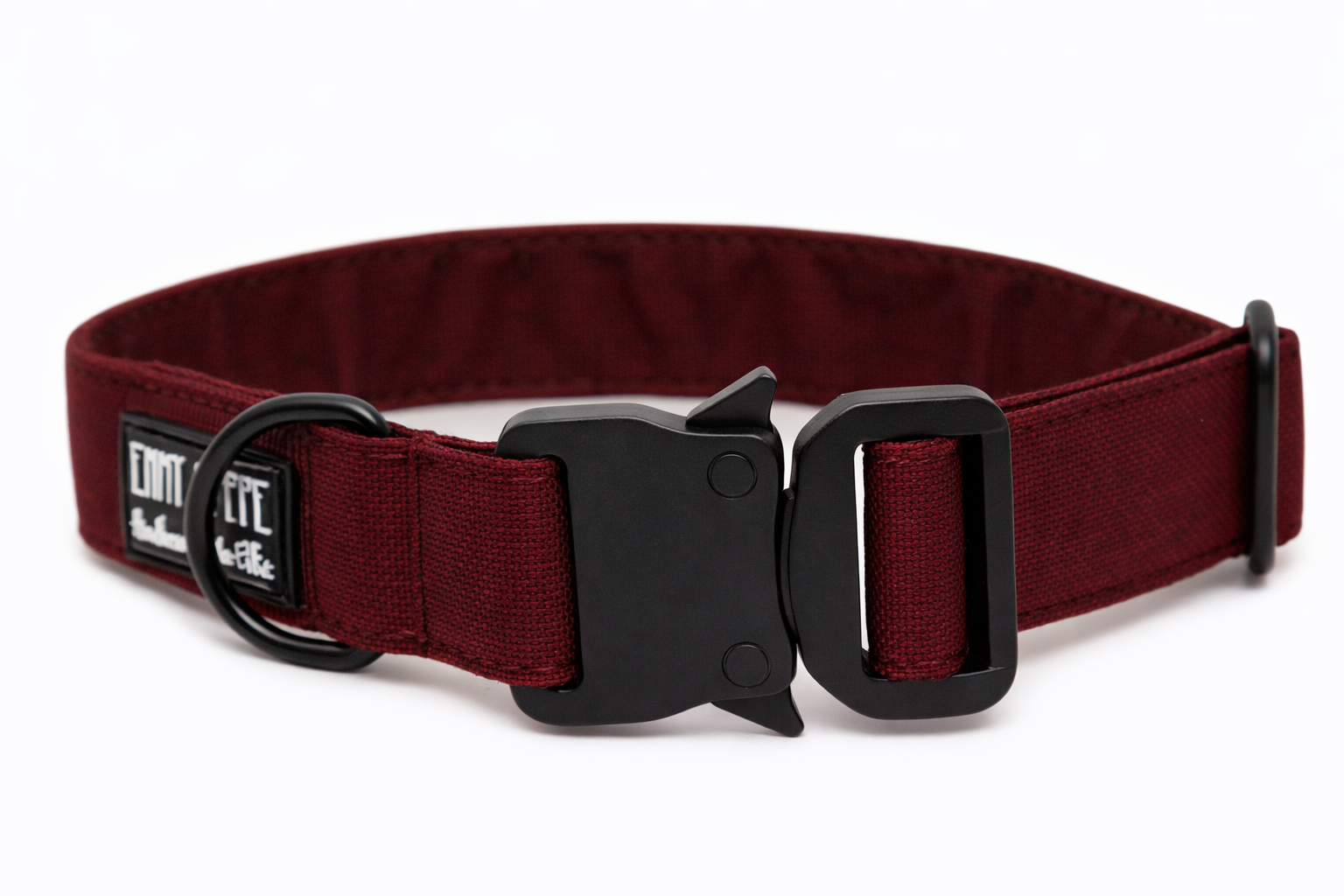 Bordeauxfarbenes Hundehalsband aus Stoff mit schwarzem Kunststoff-Sicherheitsverschluss und schwarzem Metallring zur Befestigung einer Leine. Das Halsband ist vor weißem Hintergrund fotografiert und zeigt eine breite, verstellbare Ausführung mit gepolsterter Innenseite. Auf einem kleinen Etikett am Ring ist ein Markenlogo zu erkennen.