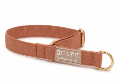 Ein rostbraunes Hundehalsband aus Stoff mit goldenen Beschlägen liegt auf weißem Grund. Auf einem beige-grauen Gummipatch steht das Logo „EMMY & PEPE“ und darunter der Slogan „Hundherum ein Wau-Effekt“.